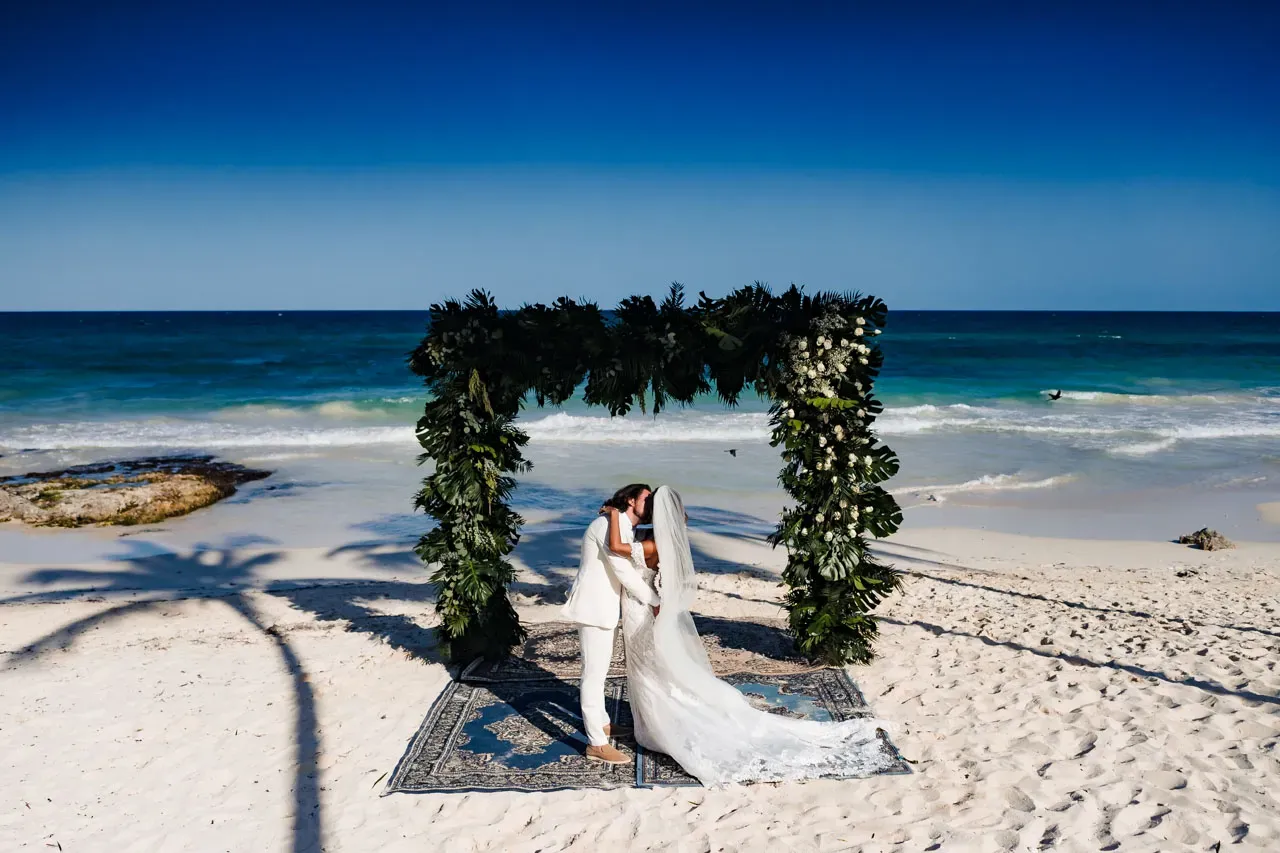 Beach wedding ceremony Cancun Riviera Maya couple kissing under tropical floral arch white sand ocean backdrop Mexico