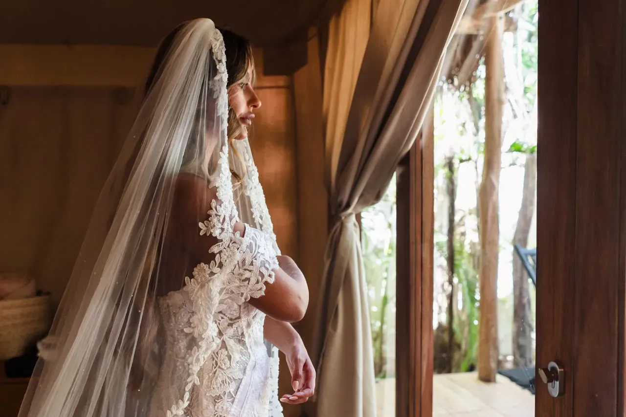 Bride in lace wedding dress and veil looking out window at tropical Riviera Maya resort before Cancun destination wedding