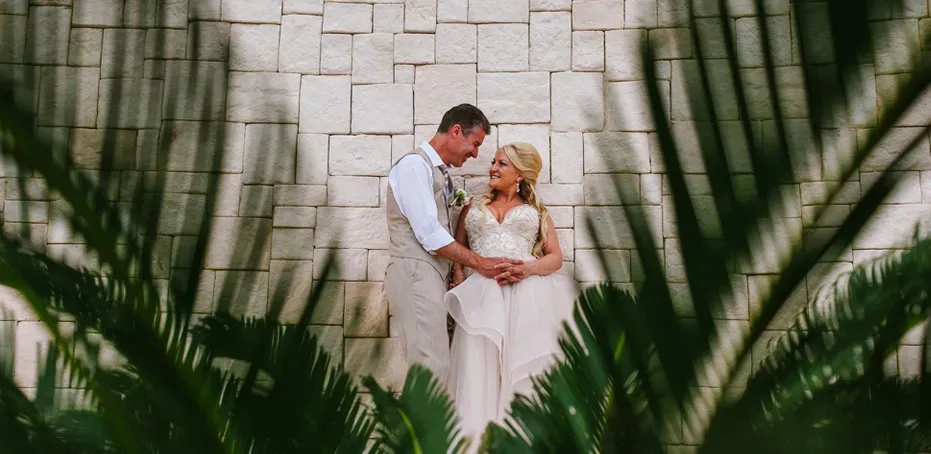 Bride and groom embrace against stone wall surrounded by tropical palms at Cancun Riviera Maya destination wedding