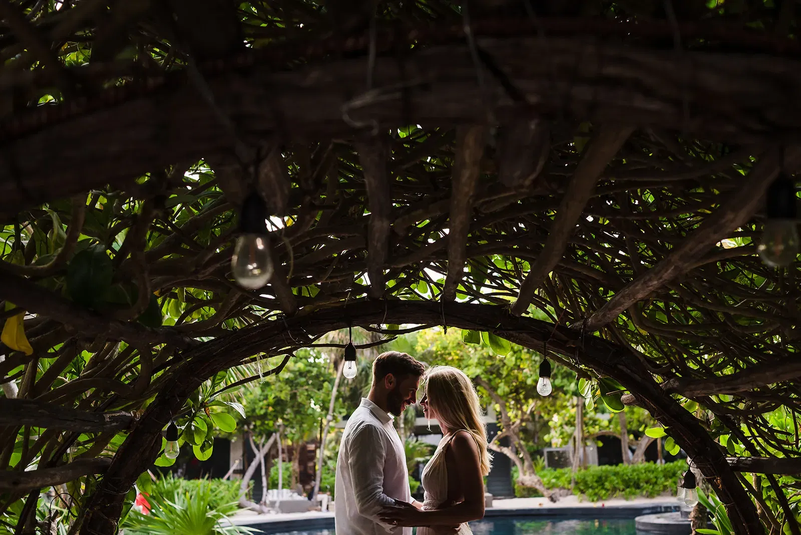 Romantic couple embracing under tropical foliage archway with string lights in Cancun Riviera Maya wedding photography