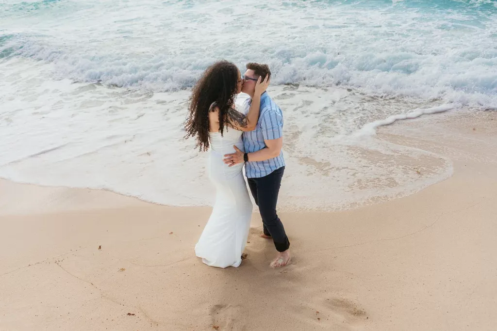 Lesbian couple kissing by the ocean barefoot in the sand.