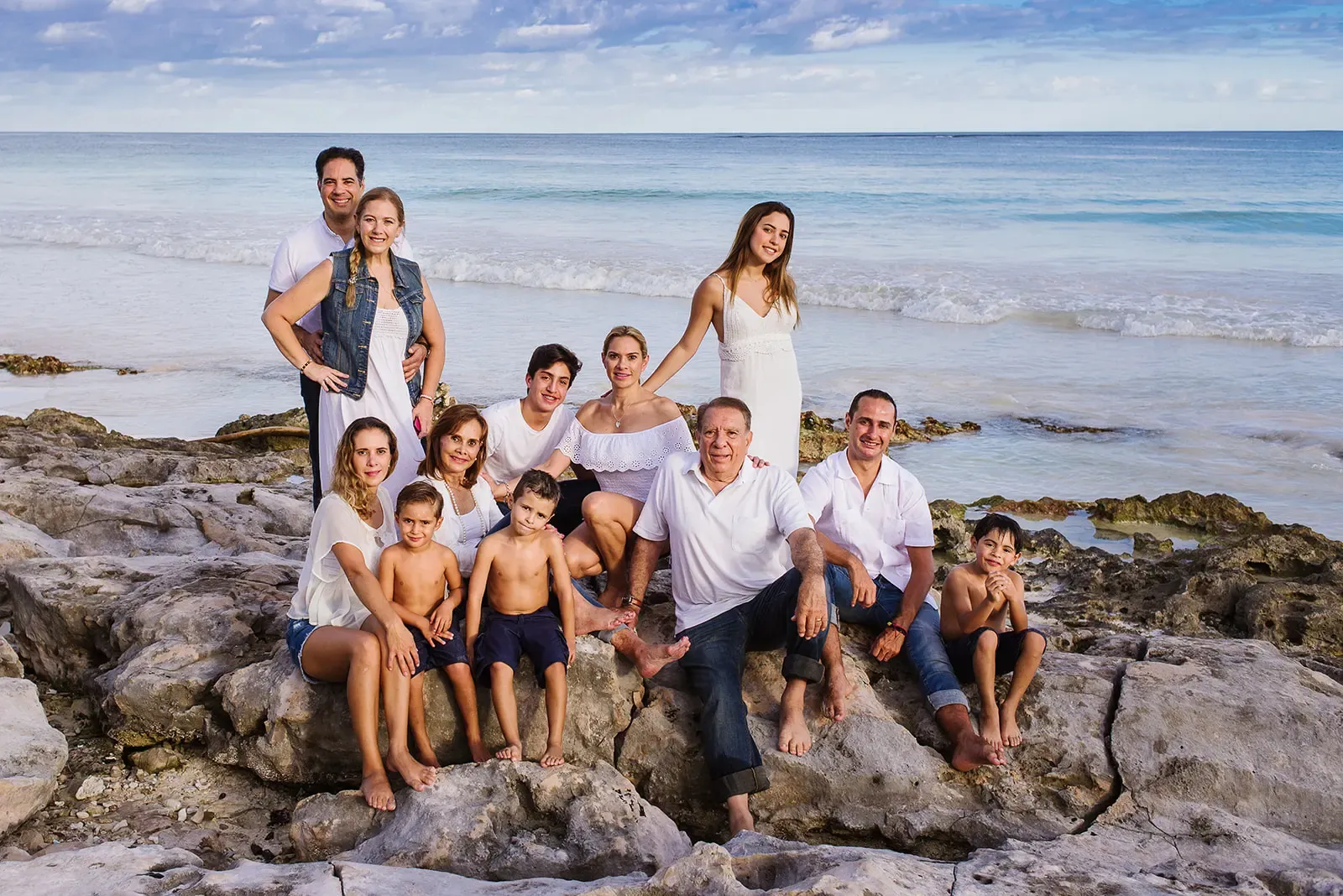 A beautiful portrait of a large, multi-generational family posing together on a rocky shoreline in Tulum. The extended family is dressed in coordinated white shirts and casual attire, smiling at the camera with the turquoise ocean waves of the Caribbean Sea creating a stunning backdrop.