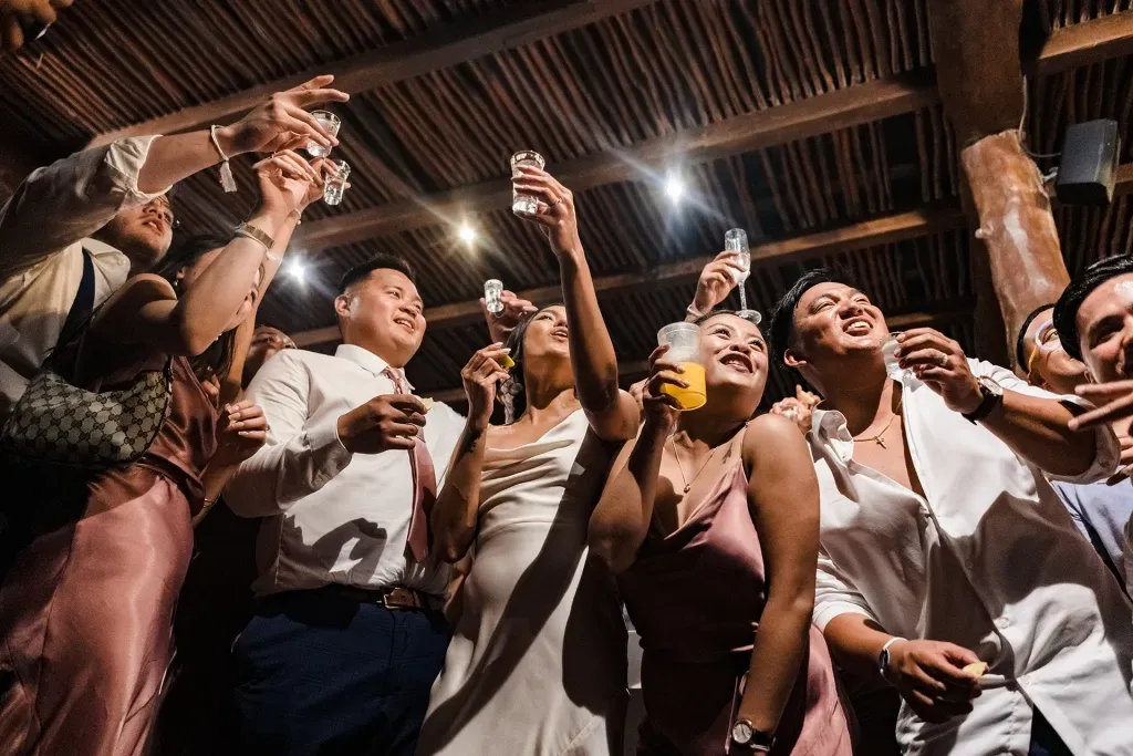 A low-angle, energetic photo of guests at a wedding reception joyfully raising their glasses for a celebratory toast. The happy group is gathered under a rustic, wooden-beamed ceiling, creating a warm and festive atmosphere.