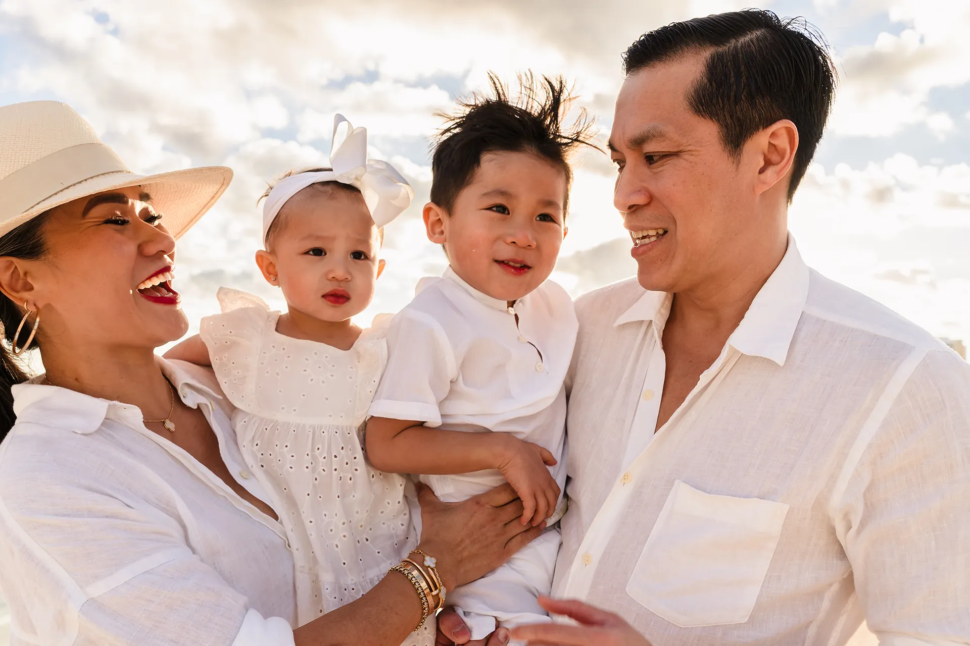 A bright, sun-kissed close-up portrait of a happy family of four. Dressed in coordinated white outfits for a beach photoshoot in Cancun, the smiling mother and father hold their young son and baby daughter in a loving embrace, creating a warm and intimate family moment.