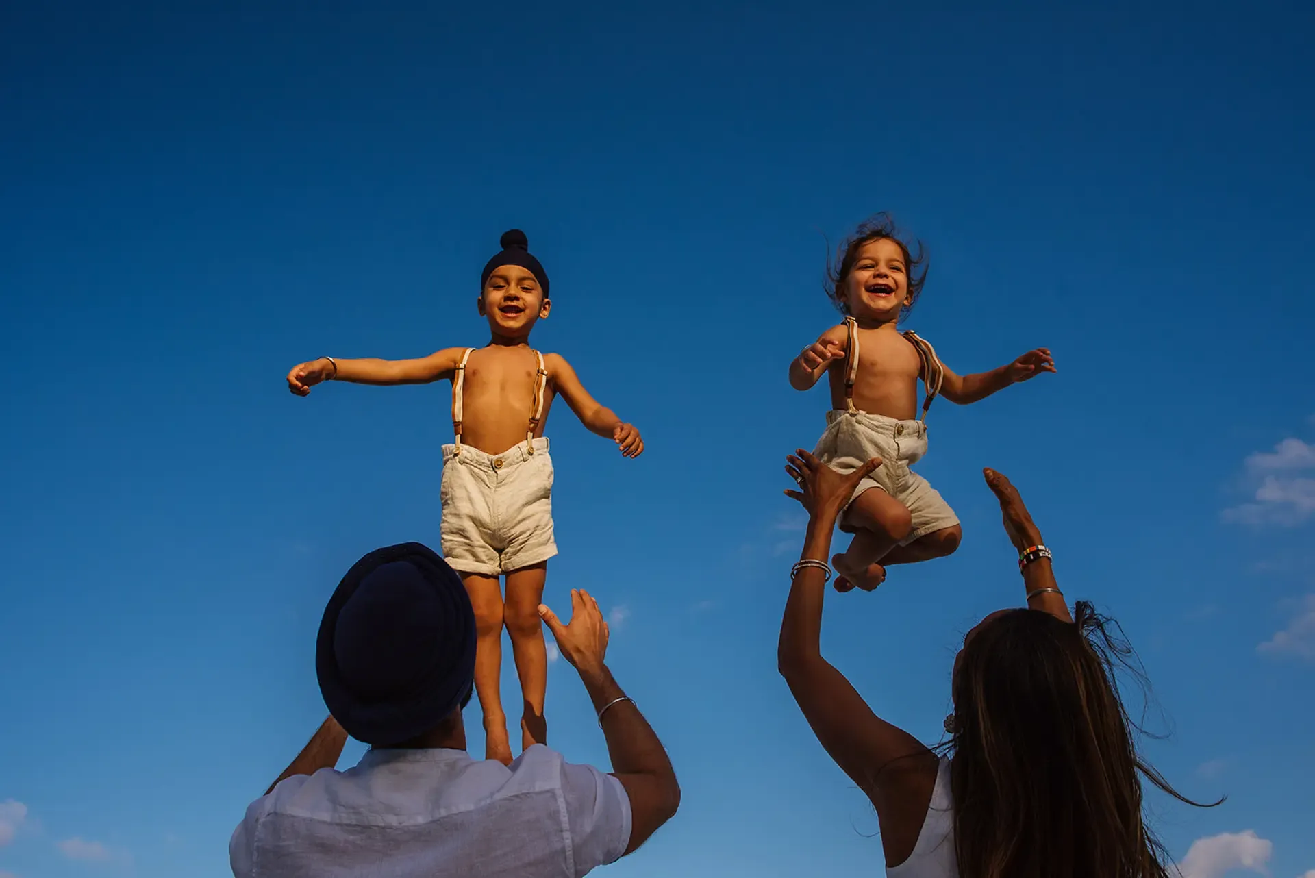 A joyful, low-angle family photograph of two young children laughing as they are playfully tossed into a clear blue sky by their parents. One child, a Sikh boy wearing a patka, and his sibling have their arms outstretched, capturing a beautiful and carefree moment of childhood bliss.
