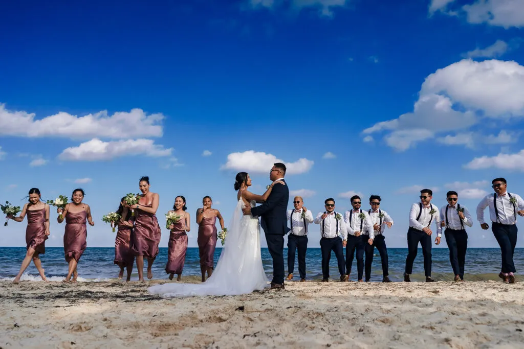 Beach wedding couple embracing on Riviera Maya sand with bridesmaids in burgundy dresses and groomsmen in suspenders
