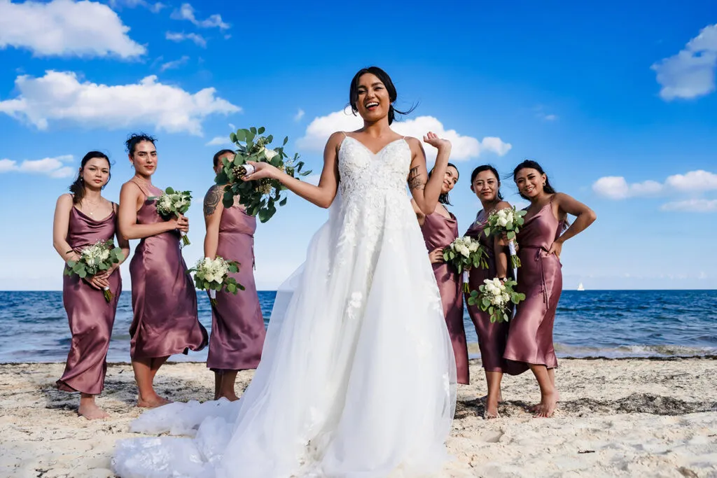 Bride and bridesmaids in dusty rose dresses pose on Cancun beach with bouquets under blue sky - Riviera Maya wedding photo