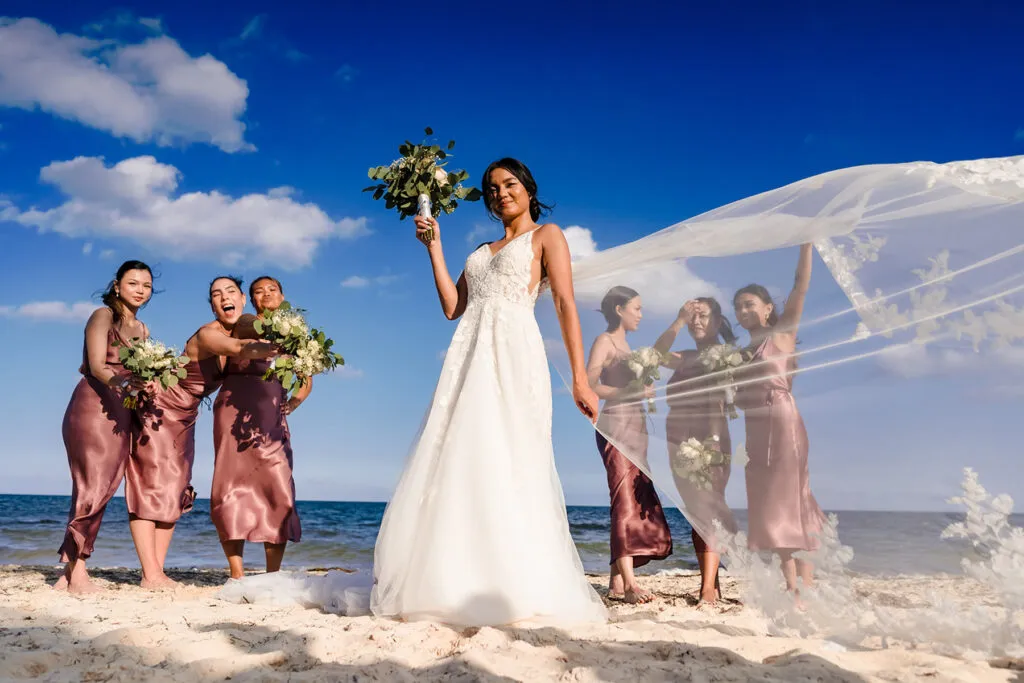 Bride with bridesmaids on Cancun beach wedding, flowing veil against blue sky - Riviera Maya destination wedding photography