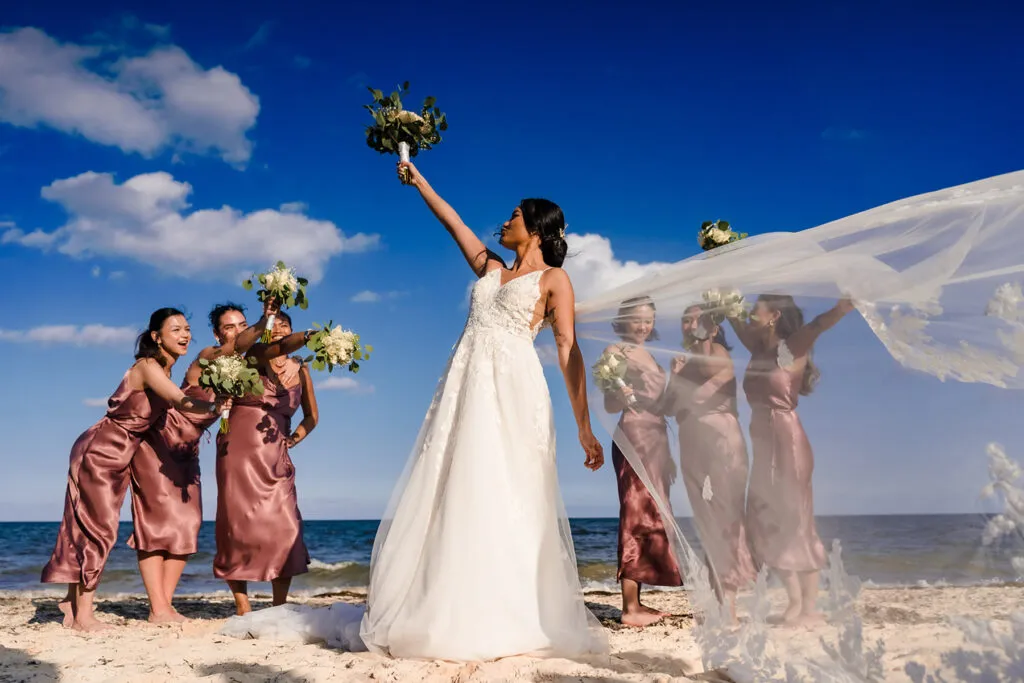 Bride tosses bouquet to bridesmaids on Cancun beach wedding with flowing veil against blue sky and ocean backdrop