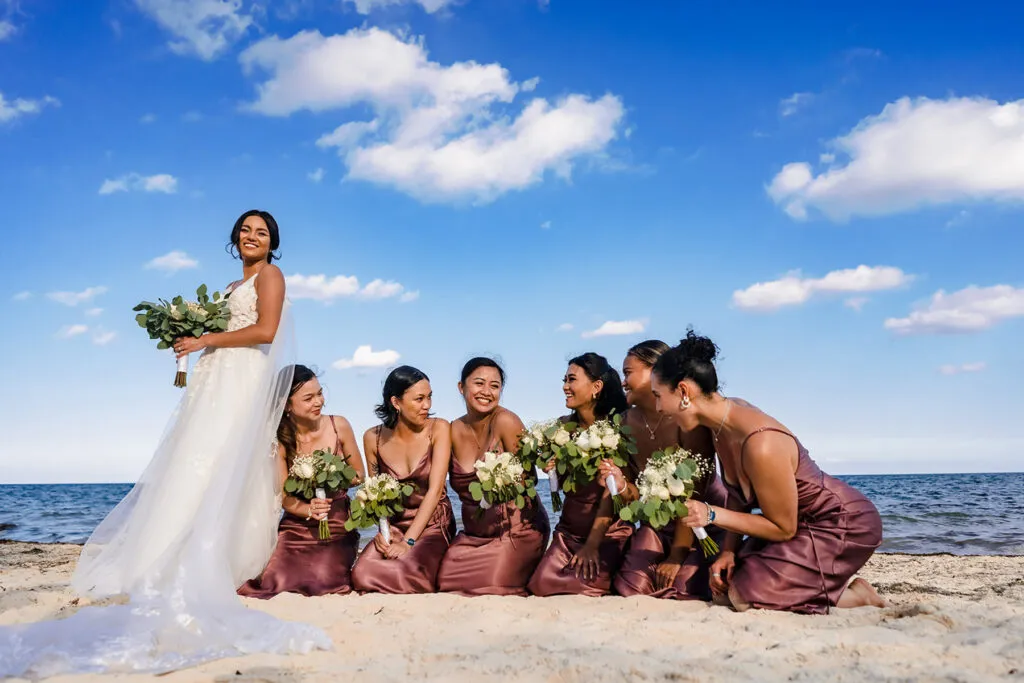 Bride with bridesmaids in burgundy dresses holding bouquets on Cancun beach wedding with blue sky and ocean backdrop