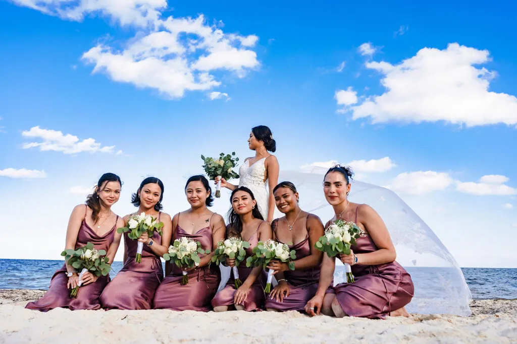 Beach wedding party in Cancun with bride and bridesmaids in mauve dresses holding white bouquets on sandy shore