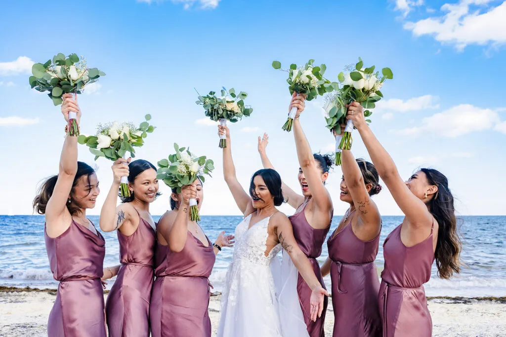 Beach wedding bridesmaids in mauve dresses celebrating with bouquets at Cancun Riviera Maya destination wedding photography