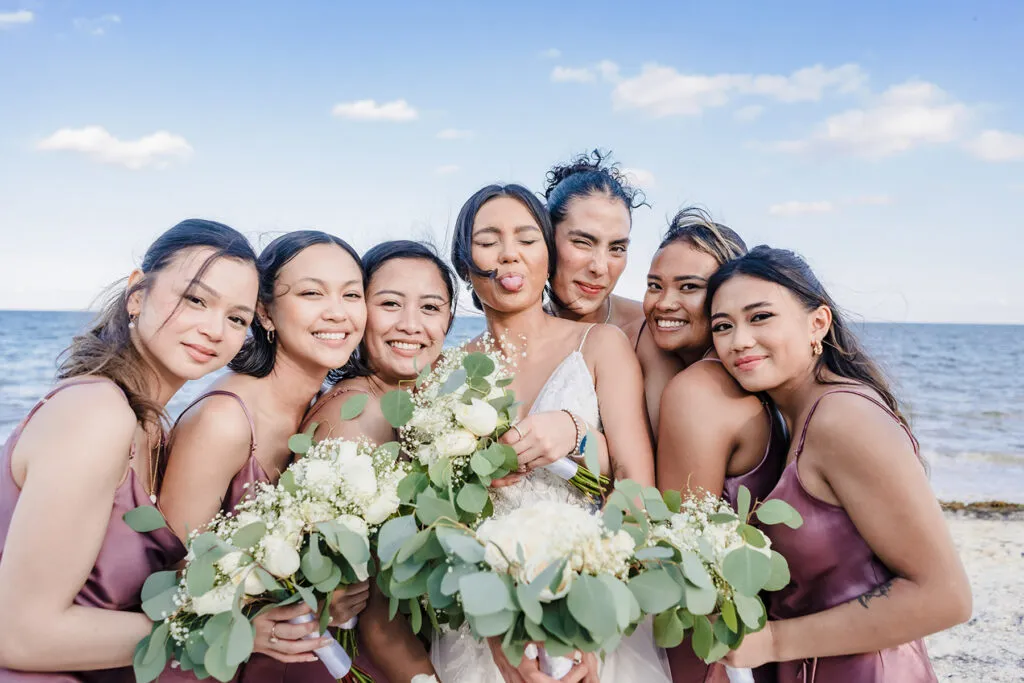 Bride with bridesmaids in mauve dresses holding white bouquets on Cancun beach - destination wedding photography Mexico