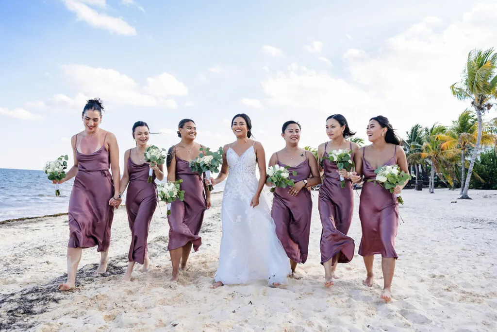 Bride with bridesmaids in purple dresses walking on Cancun beach - destination wedding photography Riviera Maya Mexico