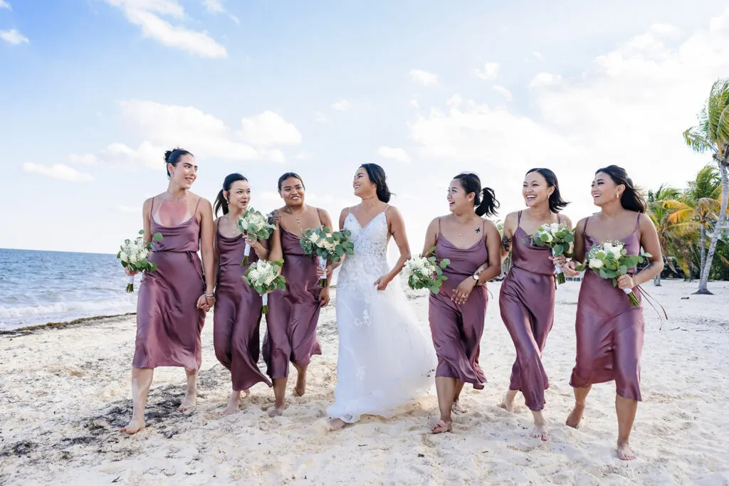 Bride and bridesmaids in mauve dresses walking on Cancun beach holding bouquets at tropical destination wedding Mexico