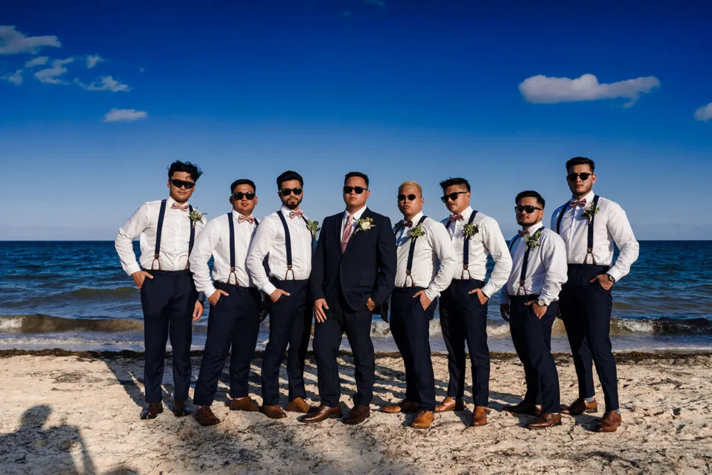 Beach wedding groomsmen in navy suits and suspenders posing on Cancun shore with ocean backdrop and blue sky