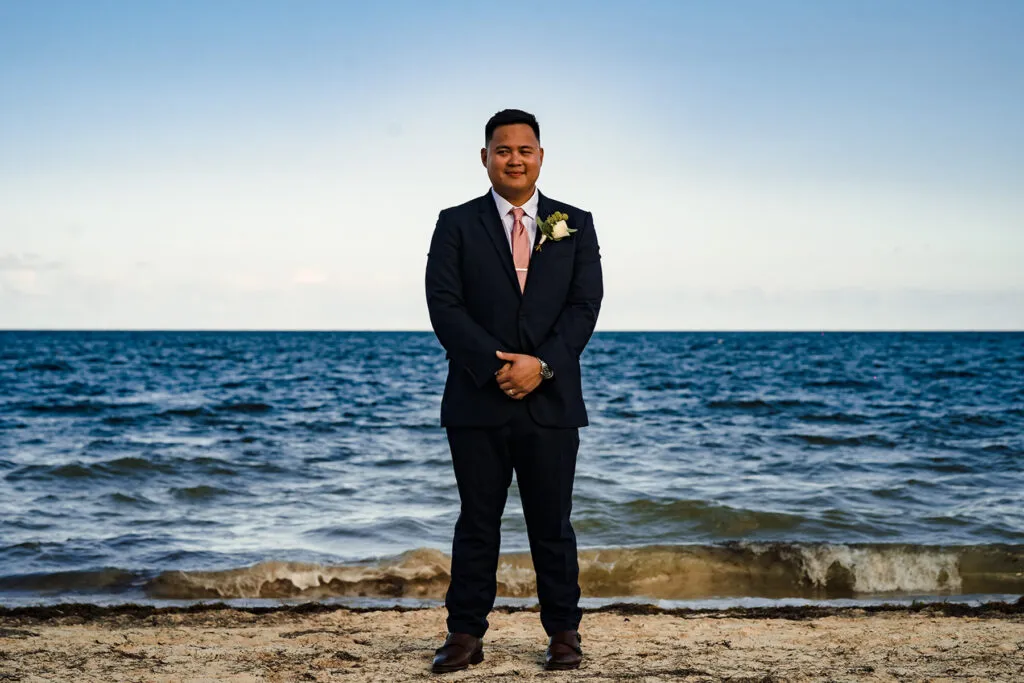 Groom in navy suit with boutonniere posing on Cancun beach during destination wedding photography session in Riviera Maya
