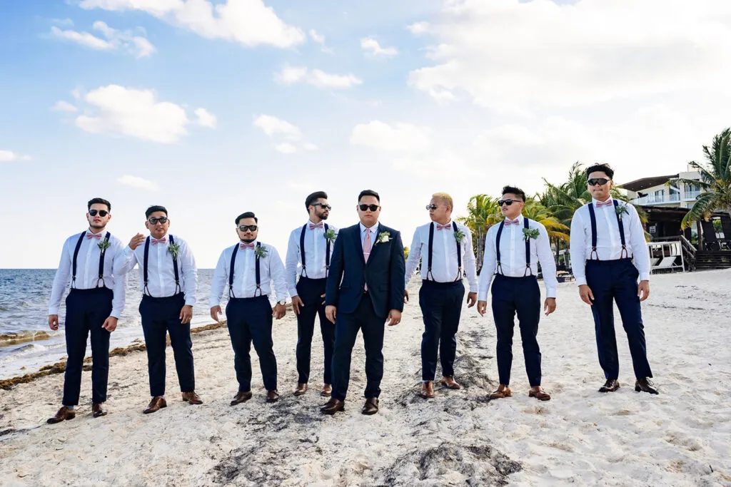 Beach wedding groomsmen in suspenders and bow ties pose on white sand beach in Cancun Riviera Maya Mexico destination wedding