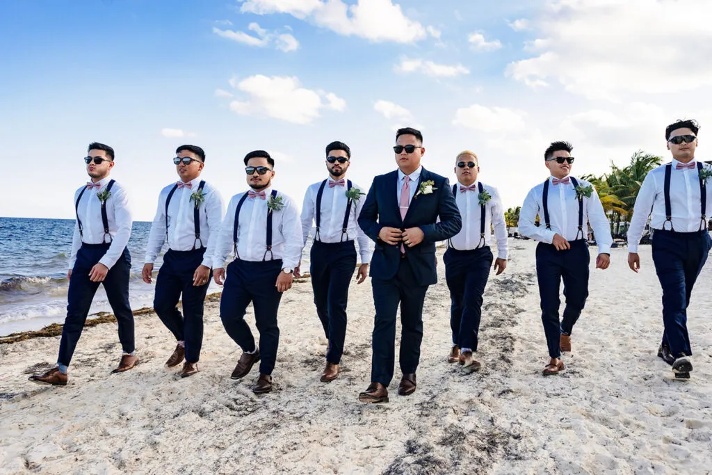 Beach wedding groomsmen walking on Cancun sand wearing sunglasses, suspenders and boutonnieres with ocean backdrop