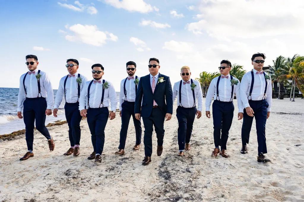Beach wedding groomsmen in suspenders and bow ties walking on Cancun sand with palm trees and Caribbean Sea backdrop