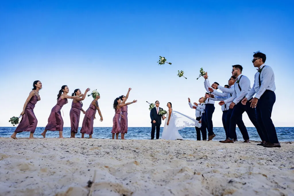 Beach wedding party throwing bouquets on Riviera Maya sand with bride groom bridesmaids groomsmen under blue sky Mexico