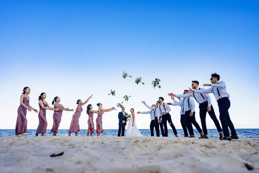 Beach wedding party throwing bouquets at bride and groom on Cancun sand with blue sky and Caribbean Sea backdrop