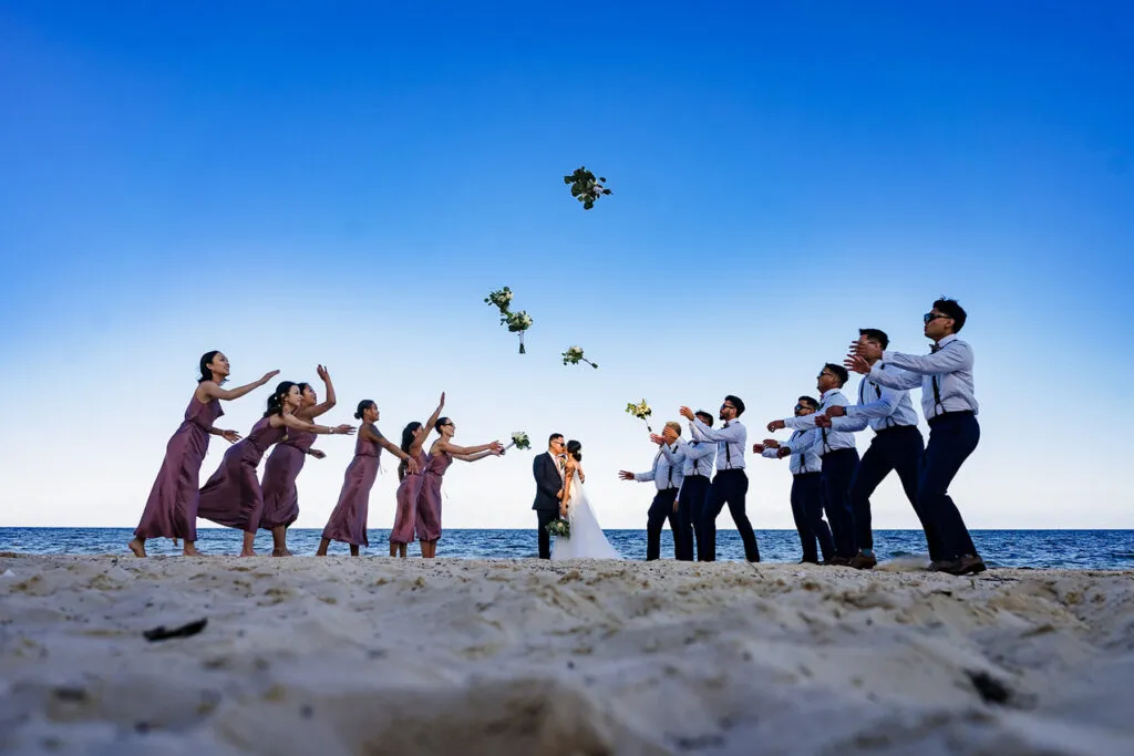 Beach wedding party throwing bouquets on Riviera Maya sand with blue ocean and sky backdrop in Cancun Mexico