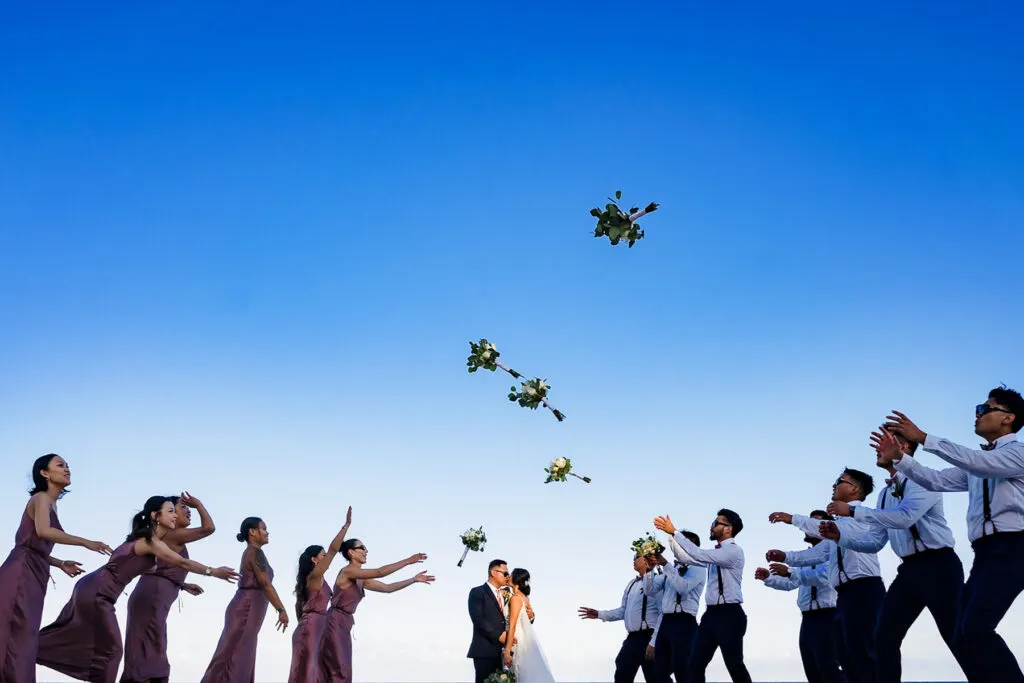 Bride and groom kissing as wedding party throws bouquets in air under blue Riviera Maya sky during destination wedding