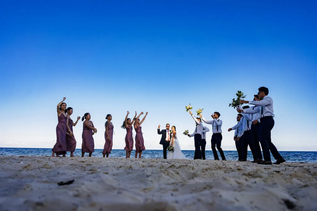 Beach wedding celebration in Cancun with bride, groom and wedding party cheering on sandy shore under blue sky