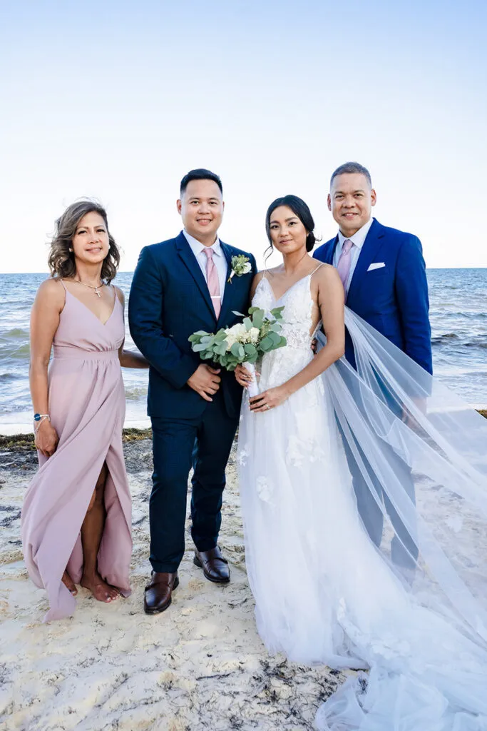 Beach wedding family portrait in Cancun with bride, groom and parents on white sand overlooking turquoise Caribbean Sea