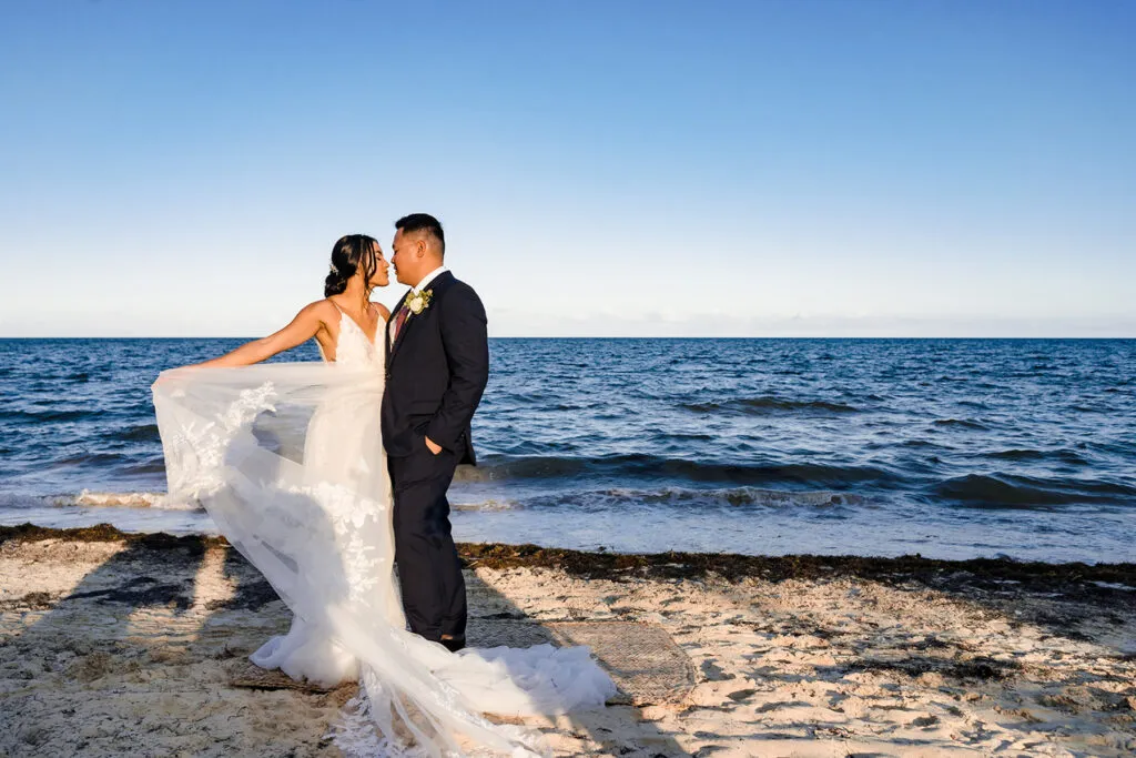 Beach wedding couple embracing at sunset in Cancun Mexico with flowing white dress and ocean waves backdrop