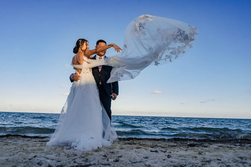 Bride and groom embracing on Cancun beach with flowing veil at sunset - romantic Riviera Maya wedding photography