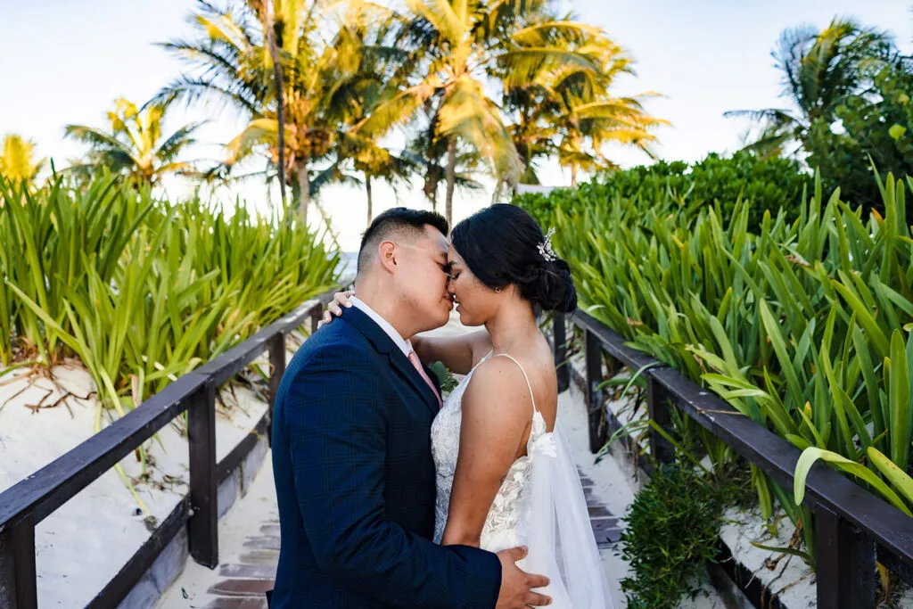Romantic newlyweds kissing on tropical boardwalk surrounded by palm trees in Cancun Riviera Maya destination wedding