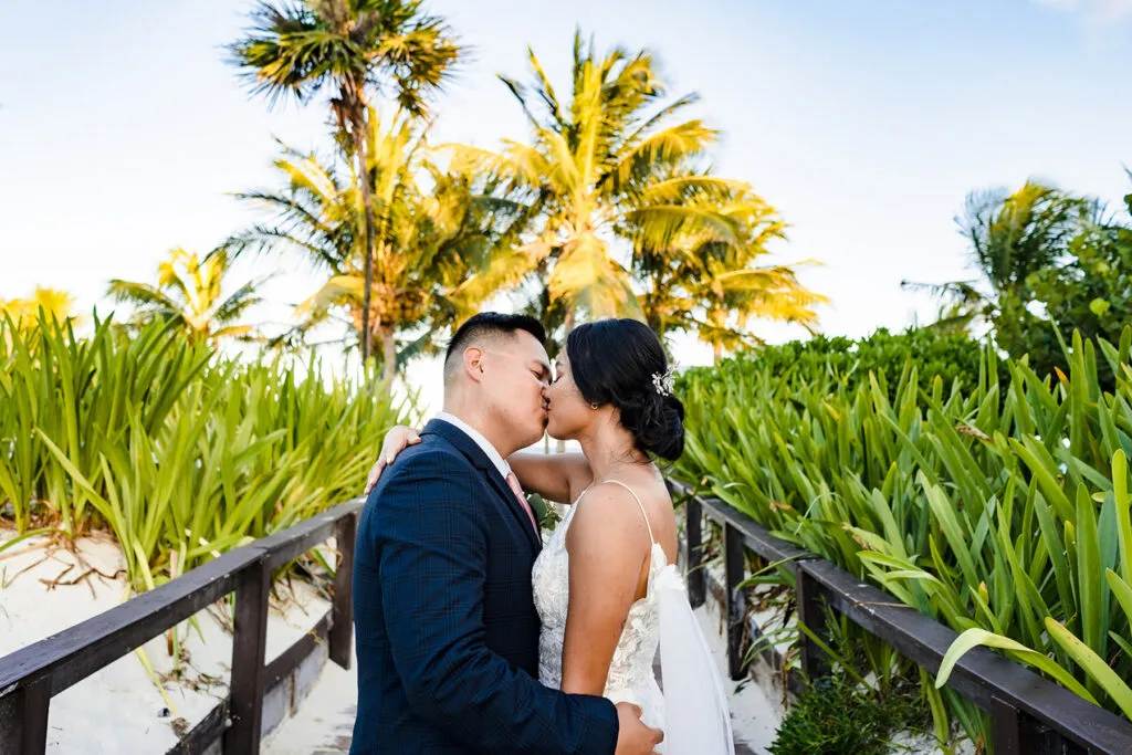 Bride and groom kissing on tropical boardwalk surrounded by palm trees in Cancun Riviera Maya destination wedding photography