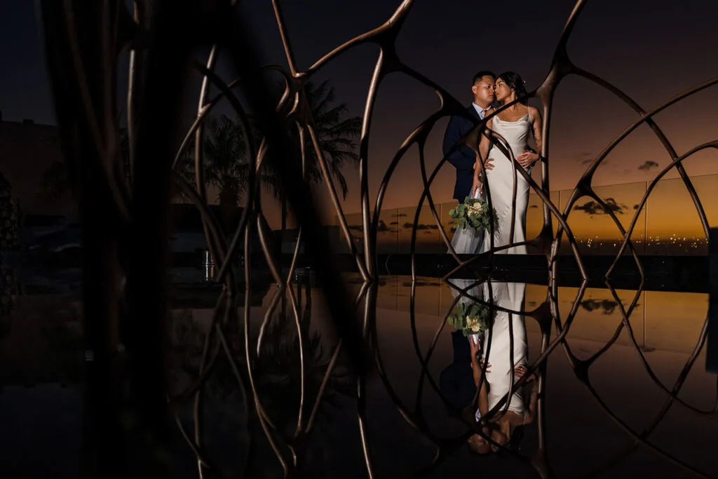 Romantic wedding couple portrait at sunset in Cancun with modern sculpture and palm trees reflected in water