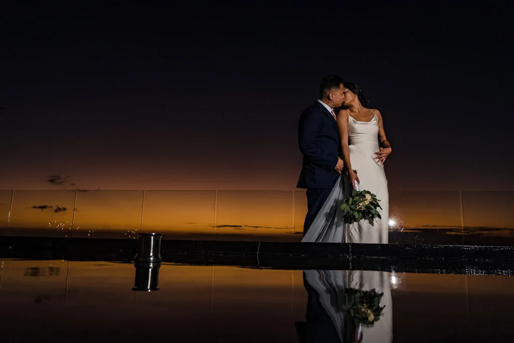 Romantic sunset wedding portrait of bride and groom kissing in Cancun with dramatic sky reflection on water