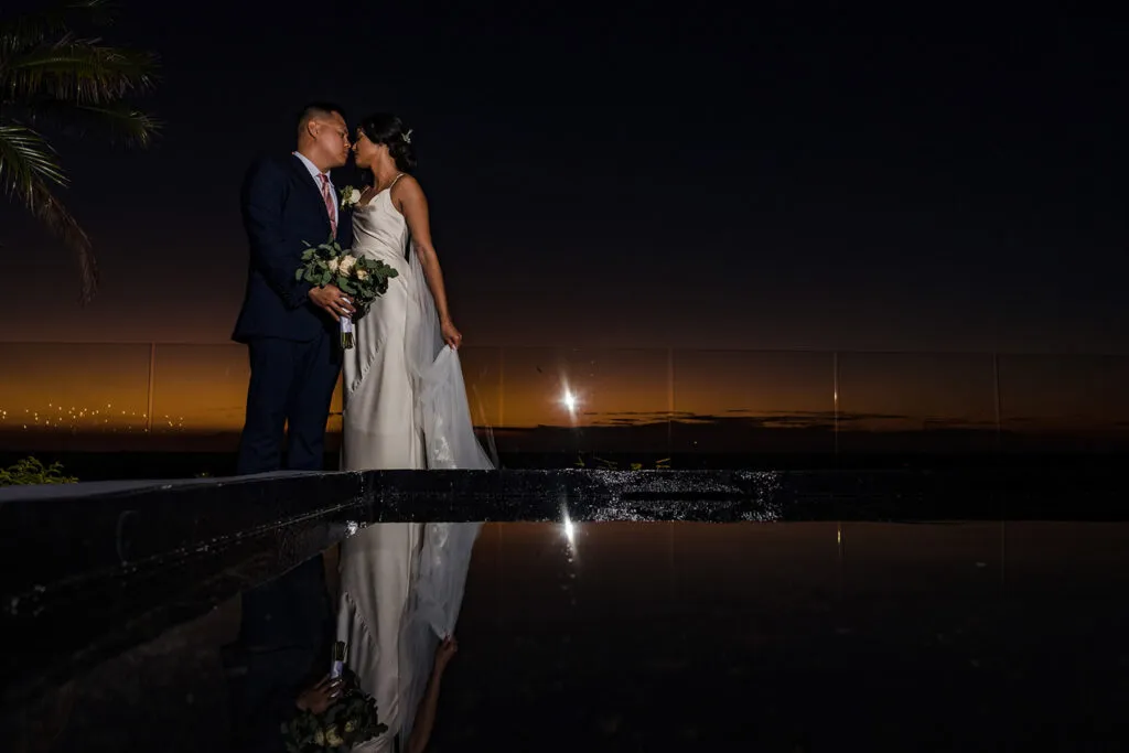 Romantic sunset wedding couple kissing by pool in Cancun Mexico with palm trees and golden sky reflection