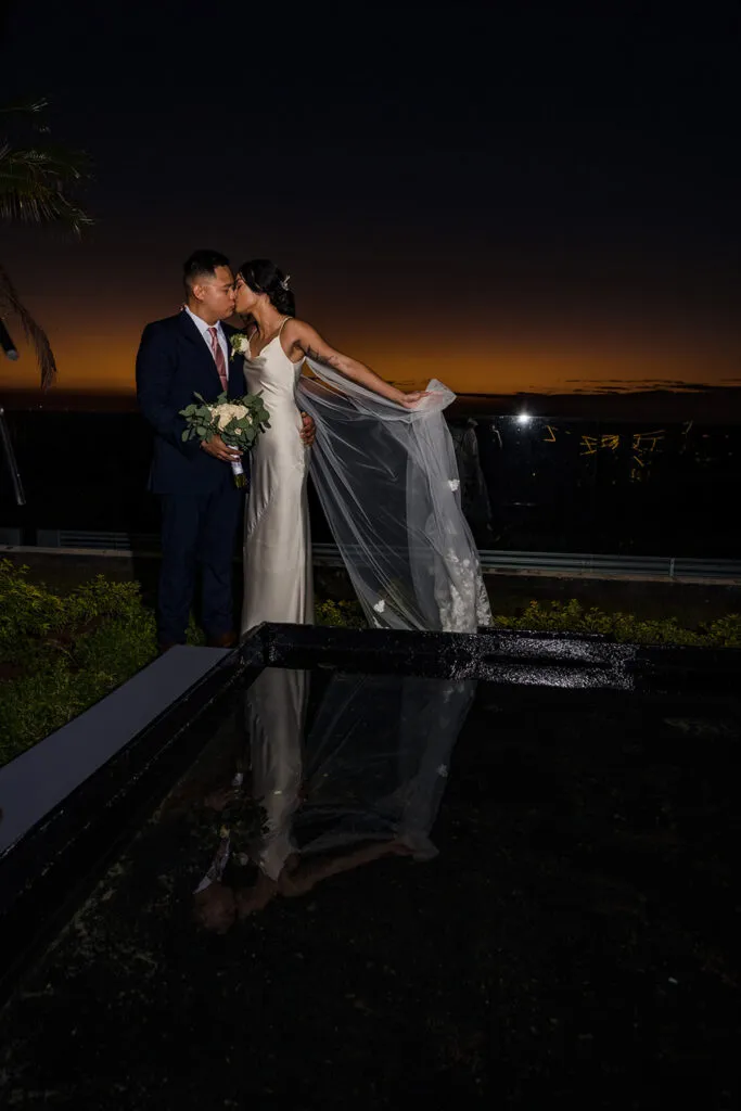 Romantic sunset wedding portrait of couple kissing by pool in Cancun, Mexico with bride's veil flowing in breeze