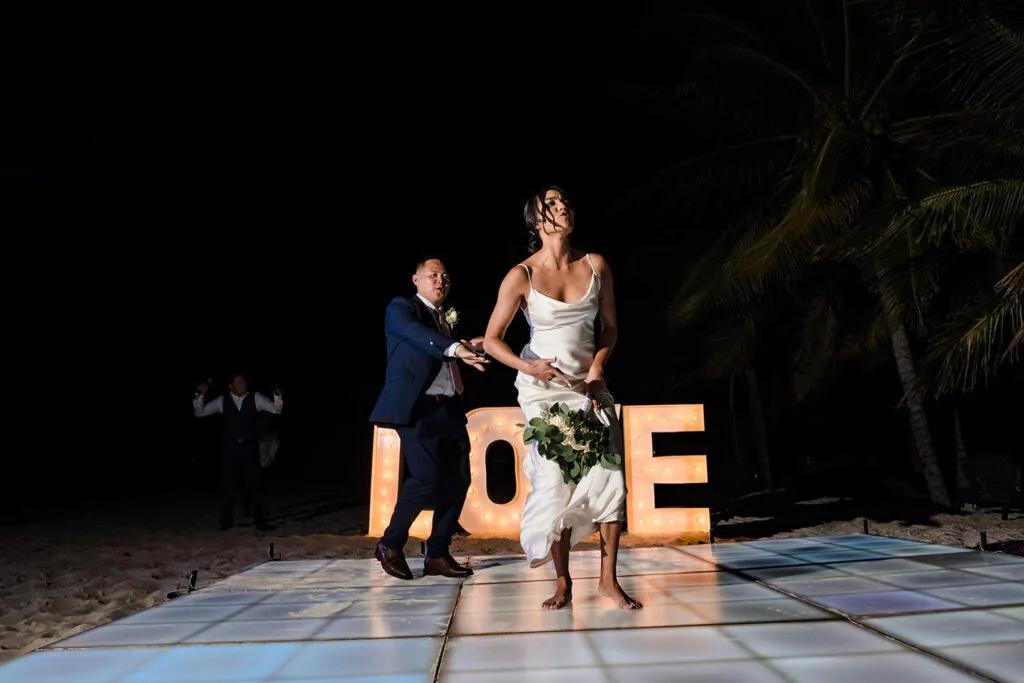Bride and groom dancing at night on beach in Cancun Mexico with illuminated LOVE sign and palm trees at destination wedding