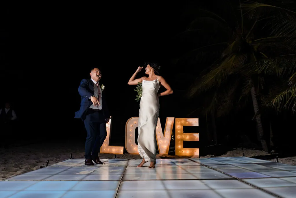 Bride and groom celebrating with tequila shots beside illuminated LOVE sign on beach dance floor in Cancun wedding photography