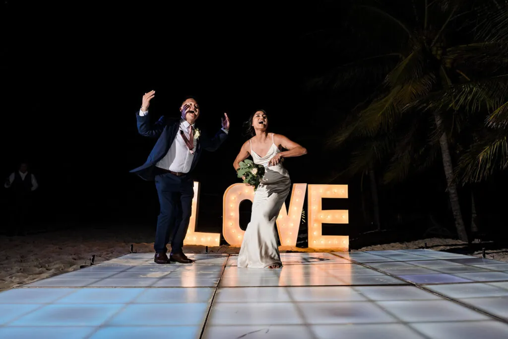 Bride and groom celebrating at night beach wedding reception in Cancun with illuminated LOVE sign and palm trees