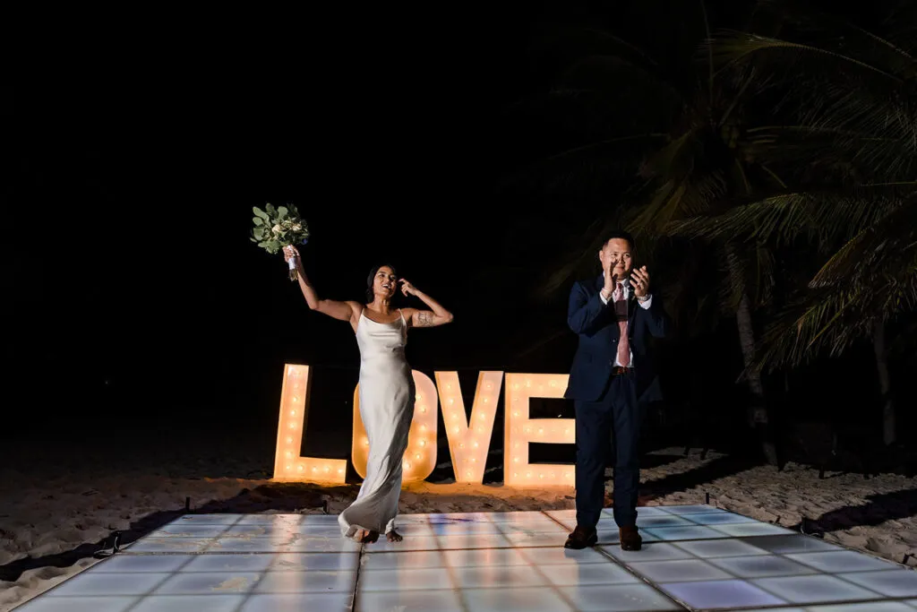 Newlyweds celebrating on illuminated dance floor with LOVE marquee sign at romantic Cancun beach wedding reception