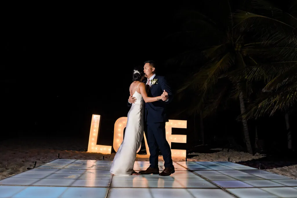 Beach wedding first dance in Cancun with illuminated LOVE sign and palm trees at night - Riviera Maya wedding photography