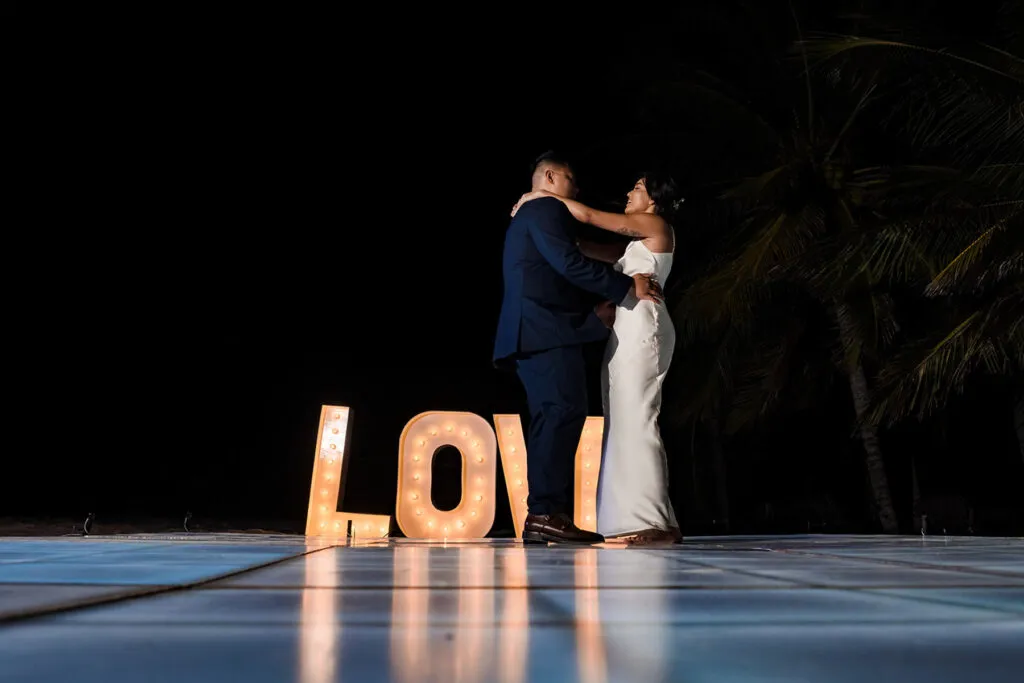 Romantic couple embracing at night wedding in Cancun with illuminated LOVE letters and palm trees in Riviera Maya Mexico