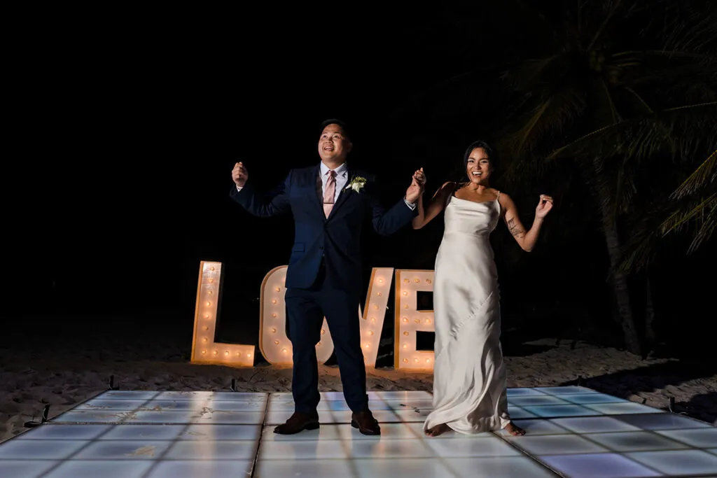 Newlyweds celebrating on beach dance floor with illuminated LOVE sign at night wedding in Cancun Riviera Maya Mexico