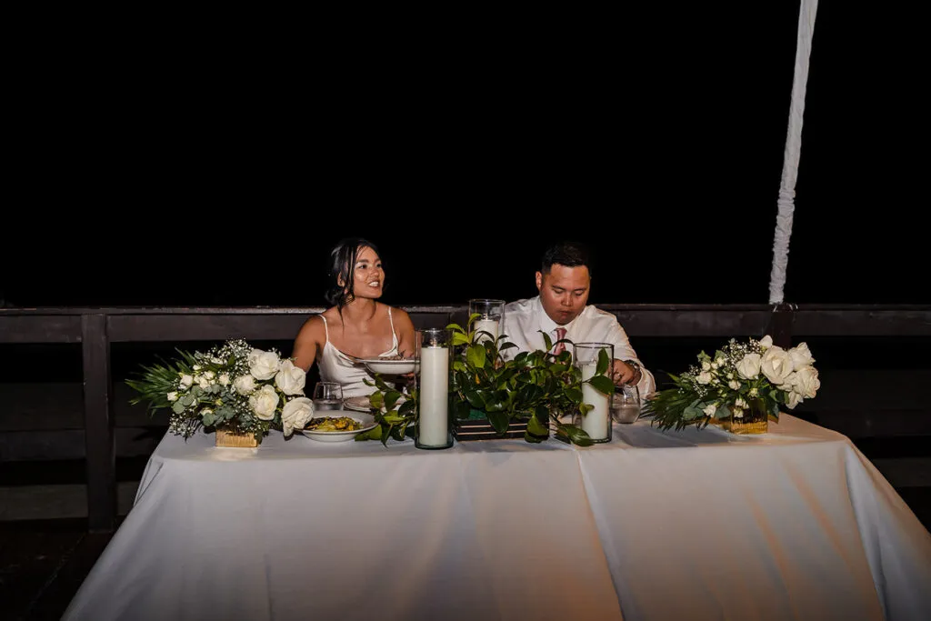Bride and groom at sweetheart table during intimate Cancun wedding reception with white floral arrangements at night