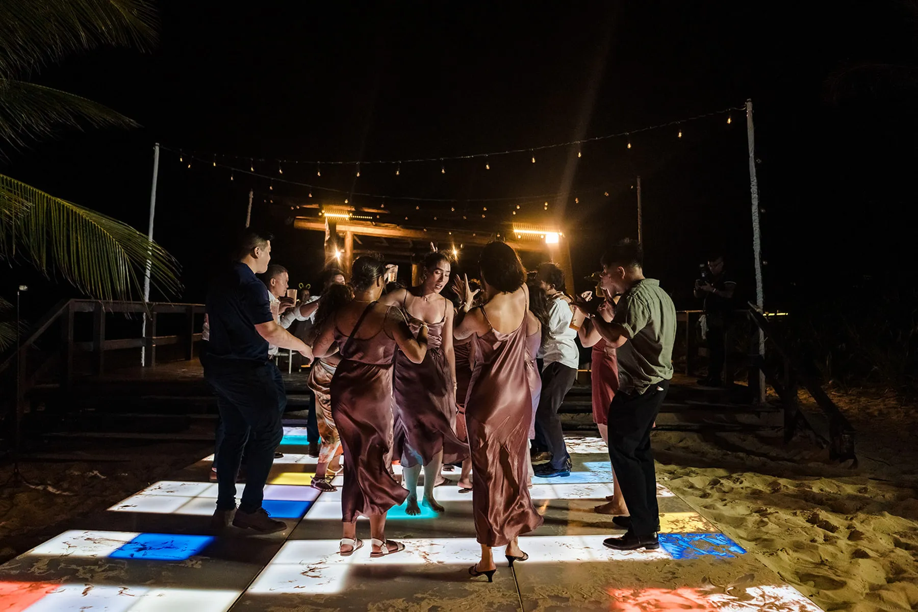 Beach wedding reception dancing under string lights in Cancun with colorful LED dance floor and tropical palm trees