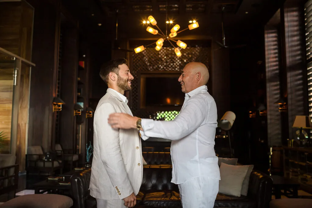Two grooms embracing in elegant white suits at upscale Cancun wedding venue with modern chandelier and luxury decor
