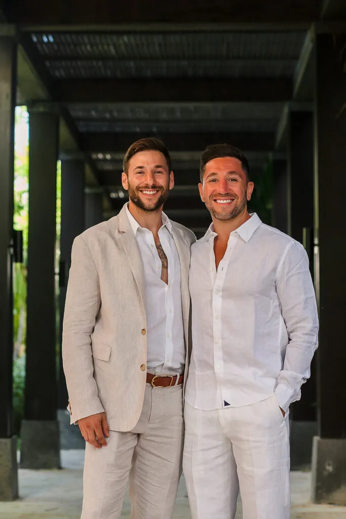 Two grooms in white linen suits smiling at luxury Cancun resort wedding venue with modern architecture backdrop