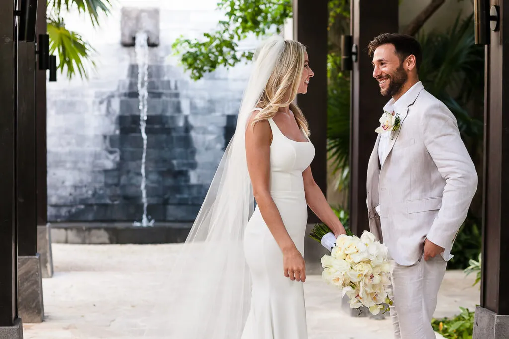 Bride and groom smiling at each other during wedding ceremony in Cancun Mexico with tropical plants and fountain backdrop